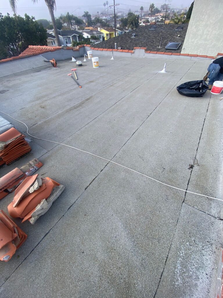 Flat residential roof in Southern California during repair work, showing gray rolled roofing surface with visible seams, vent pipes sealed with white flashing, roofing tools and buckets on the roof, stacks of clay tiles along the edge, and a neighborhood with palm trees and homes in the background.