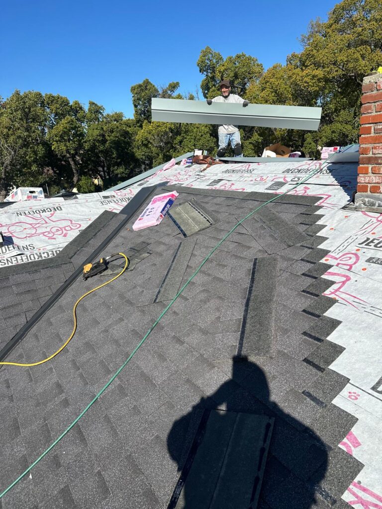 Roofer installing ridge vent and asphalt shingles during a residential roof repair project with underlayment and flashing visible.