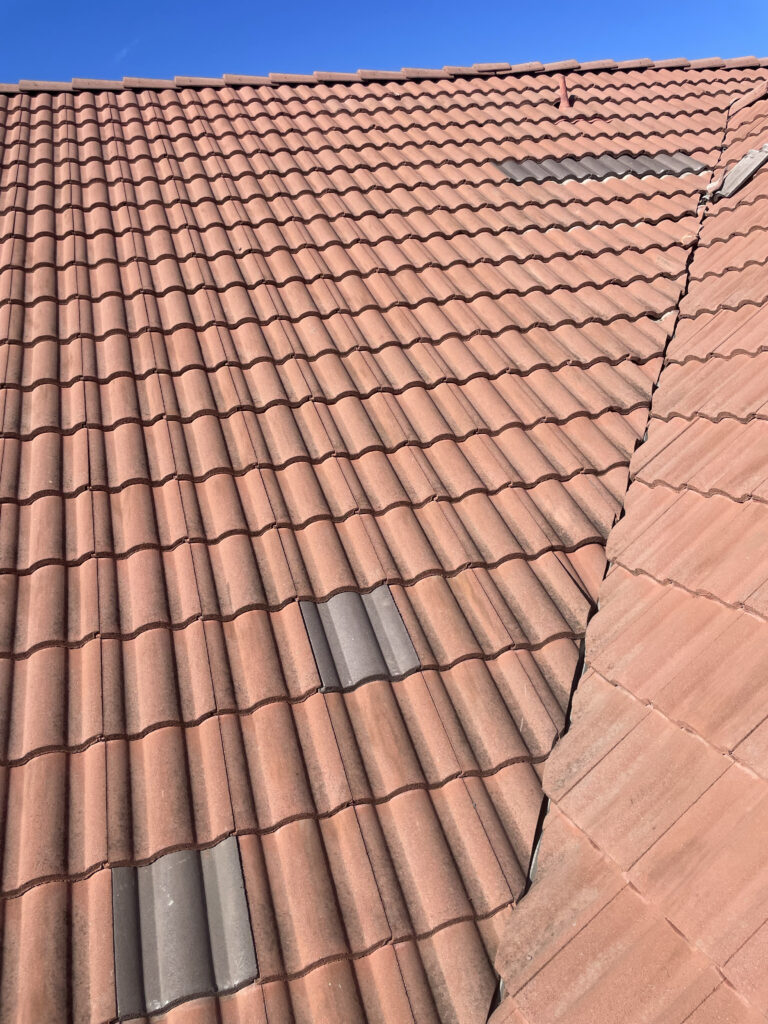 Clay tile roof on a Southern California home with a few darker replacement tiles visible, showing the curved tile pattern and roof valley under a clear blue sky.