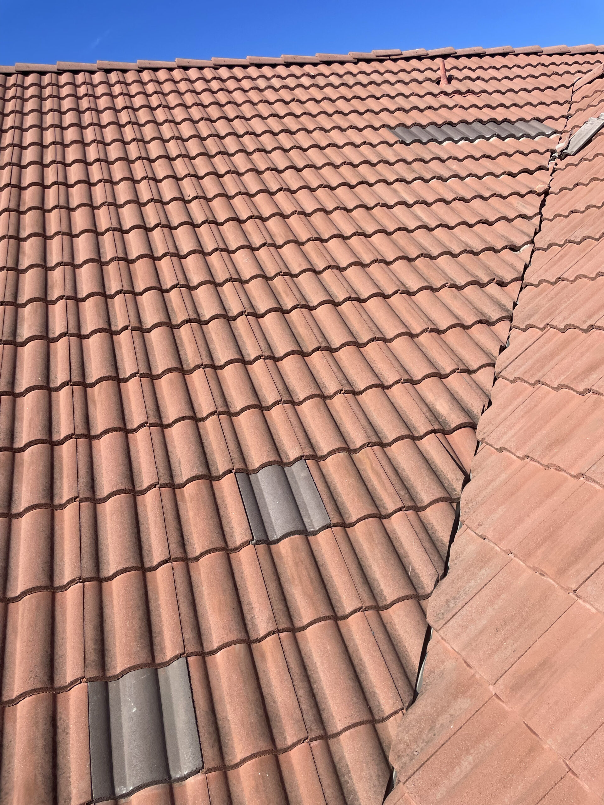 Clay tile roof on a Southern California home with a few darker replacement tiles visible, showing the curved tile pattern and roof valley under a clear blue sky.