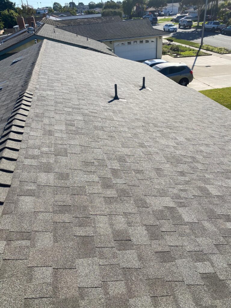 Asphalt shingle residential roof in a suburban neighborhood with two roof vent pipes and visible ridge line, photographed from the roof looking toward nearby homes and street.